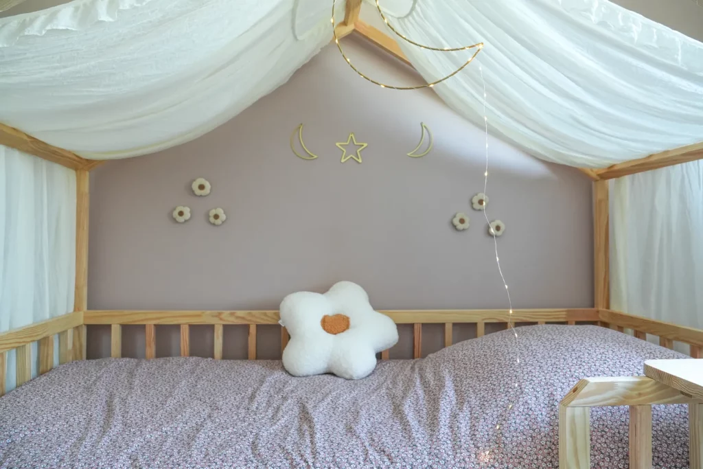 Cabane de lit artisanale en bois clair avec ciel de lit en tissu blanc, décorée de guirlandes lumineuses, d’un coussin fleur et de motifs muraux en forme de lune, étoile et fleurs. Création unique de l'Atelier de Léna.